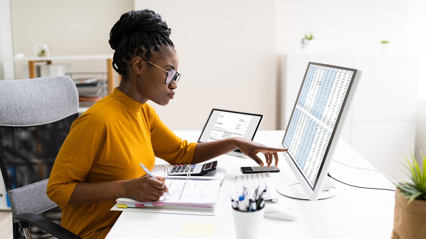 A woman sitting at her desk with an open notebook and computer monitor with a spreadsheet open.