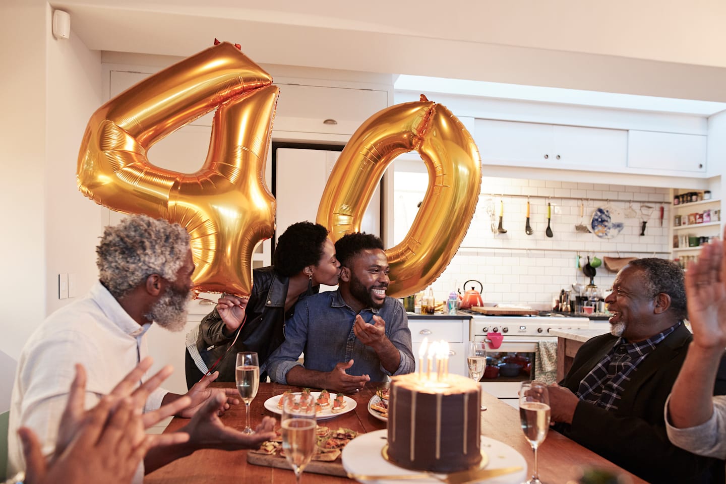 People are seated around a table celebrating a person's birthday with cake and food. Balloons in the background show a four and a zero for the birthday age of 40.