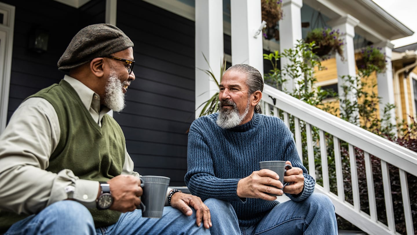 Two older men sitting on a porch drinking coffee.