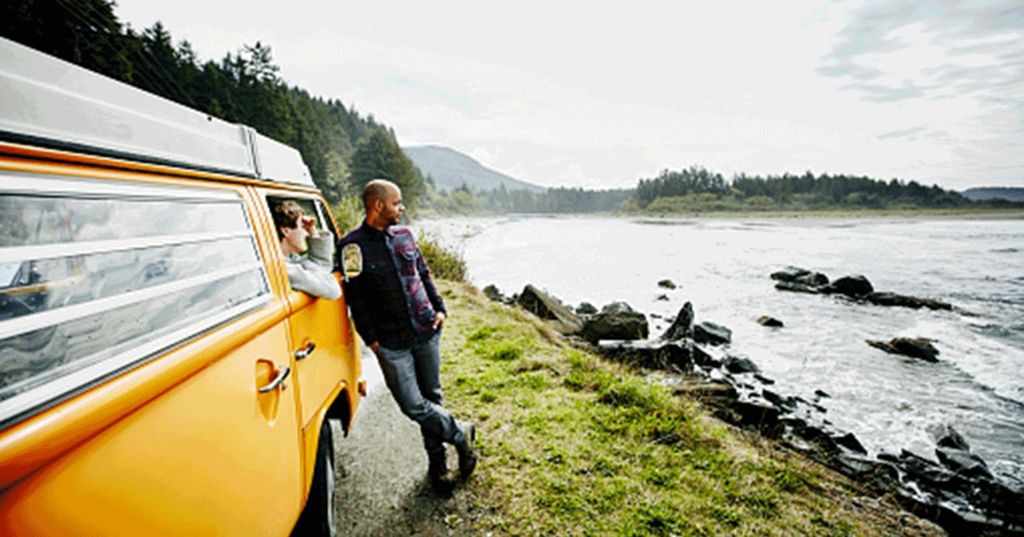 A person leaning against a yellow van with another person leaning out of the passenger window staring out into the lake.