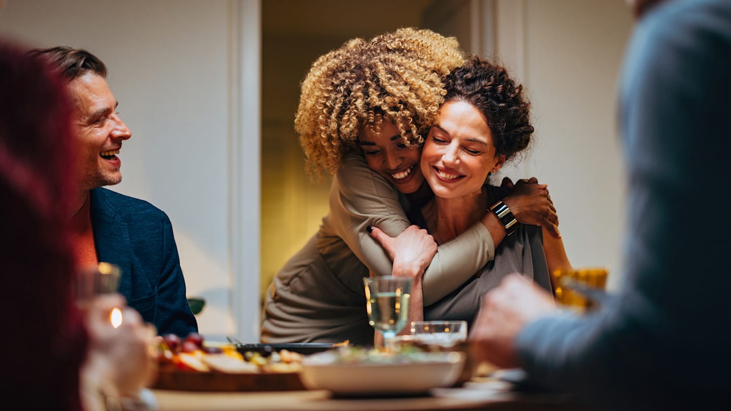 Woman hugging another woman while man watches. All are smiling and around a dining table.