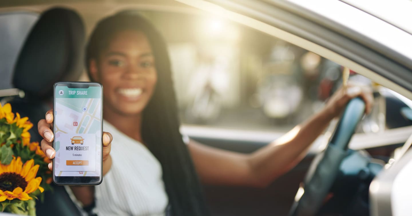 A women in a car holding up her mobile phone showing that he has a person requesting a ride share.