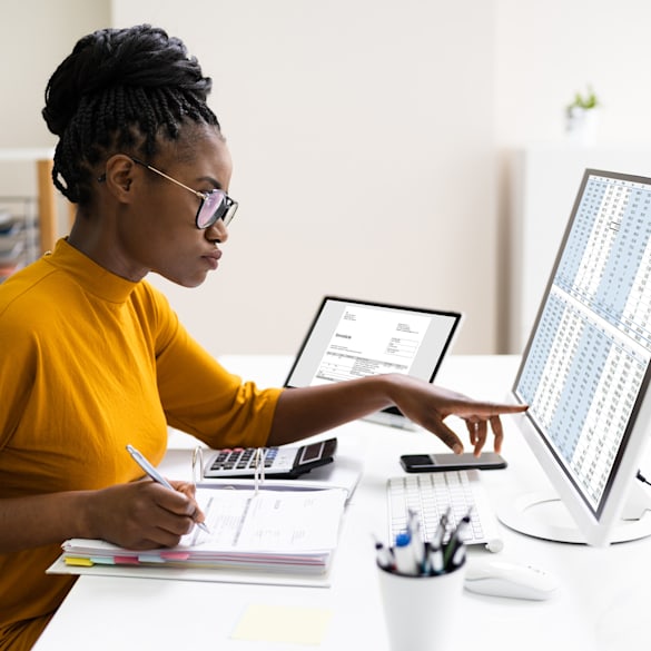 A woman sitting at her desk with an open notebook and computer monitor with a spreadsheet open.