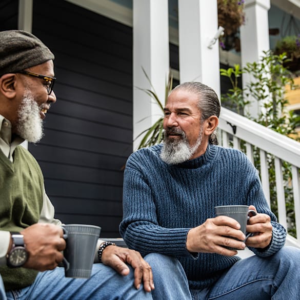 Two older men sitting on a porch drinking coffee.