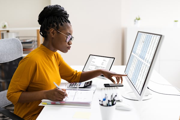 A woman sitting at her desk with an open notebook and computer monitor with a spreadsheet open.